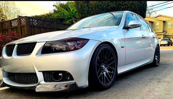 A silver sedan with dark wheels is parked on a residential street, surrounded by wooden fences, greenery, and houses in the background.