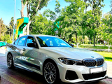 A white BMW sedan is parked at a gas station with trees in the background on a sunny day. The car's design is sleek, featuring sporty rims and a modern grille.