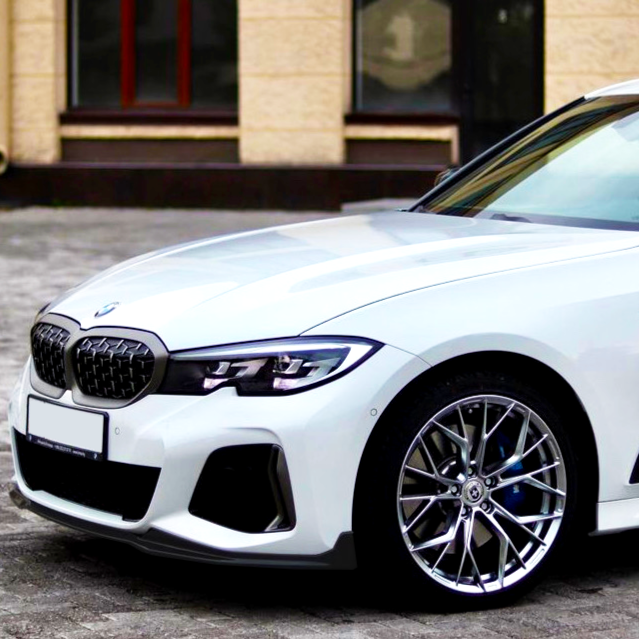 A white BMW car is parked on a cobblestone surface in front of a beige building with windows. The focus is on the front left side of the vehicle.