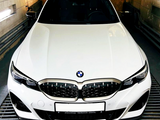 A white BMW car is stationary in an enclosed garage, with a frontal view highlighting its kidney grille. The text on the license plate reads: 