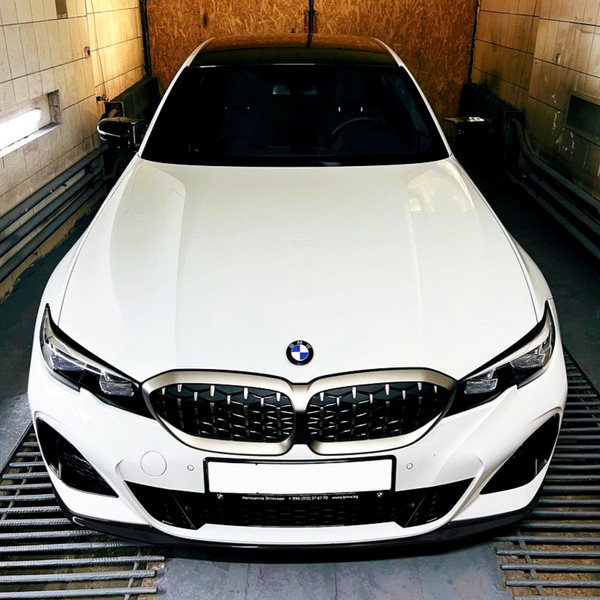 A white BMW car is stationary in an enclosed garage, with a frontal view highlighting its kidney grille. The text on the license plate reads: "Автоцентр Витаваль +996 (312) 57 77 70 www.bmkg.kg".