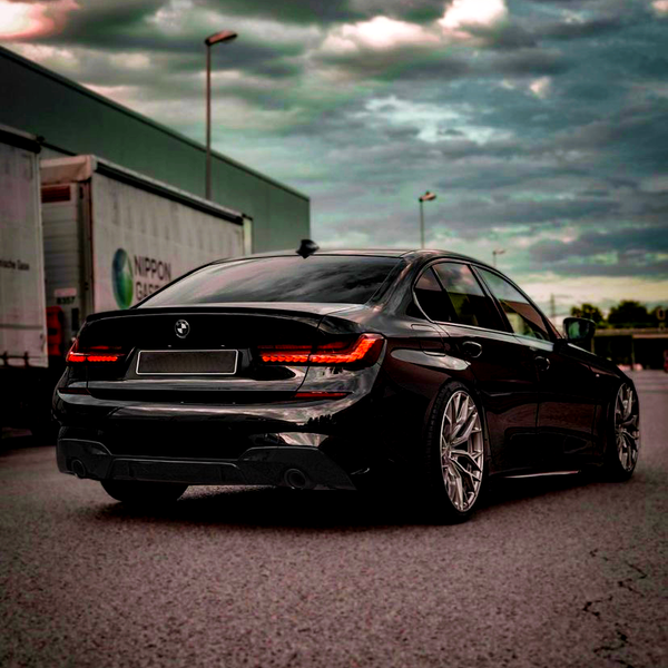 A black car with illuminated rear lights is parked on an empty street. Large wheels and a sleek design stand out. Nearby are cargo trailers under a cloudy sky.