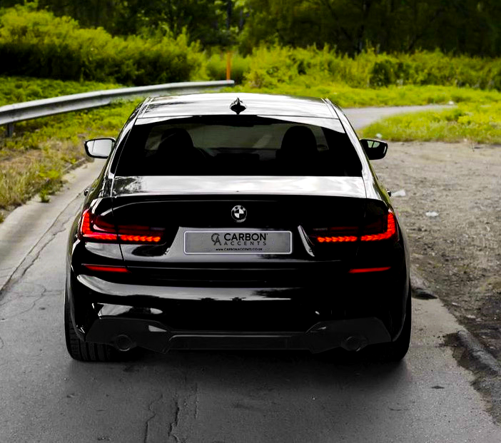 A sleek black car is parked on a paved road with greenery surrounding it. The rear license plate reads "CARBON ACCENTS" with the website "www.carbonaccents.co.uk" displayed.
