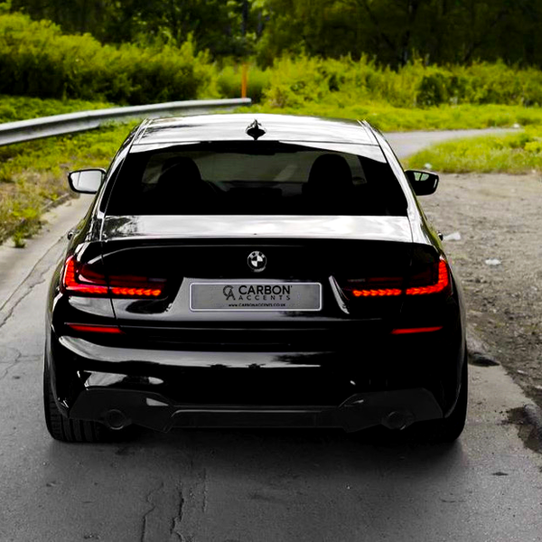 A sleek black car is parked on a paved road with greenery surrounding it. The rear license plate reads "CARBON ACCENTS" with the website "www.carbonaccents.co.uk" displayed.