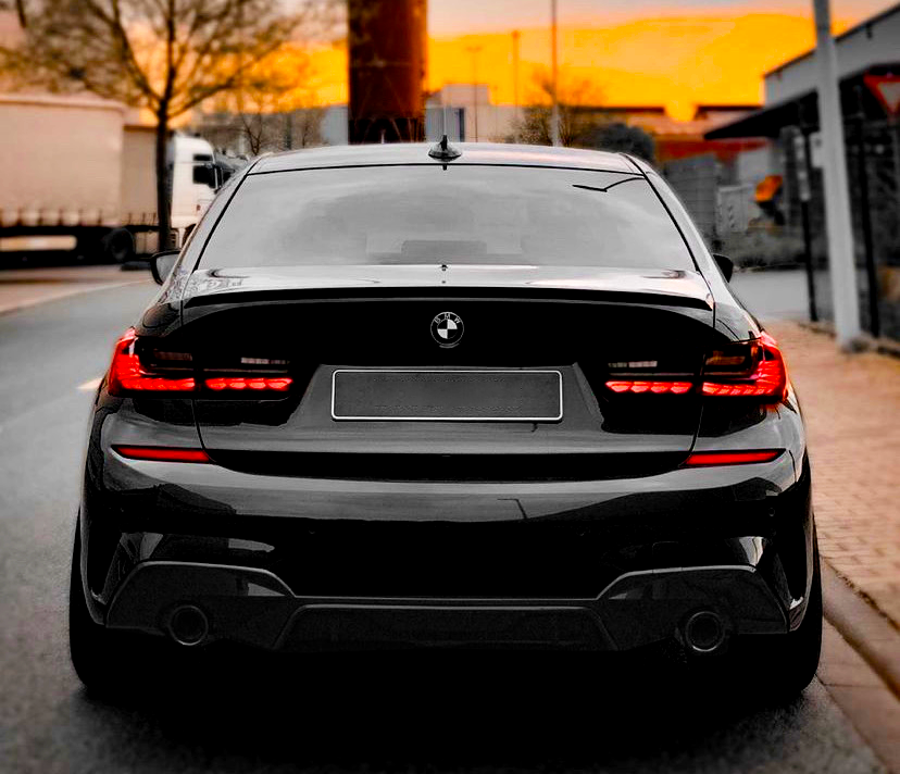 A black BMW car is parked on a street, with taillights illuminated. The sky is vibrant orange at sunset, surrounded by trees and industrial buildings.