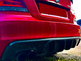 A red BMW 135i with dual exhaust pipes is parked on a grassy area. The concrete path is partially visible, and the rear of the car shows distinctive branding.