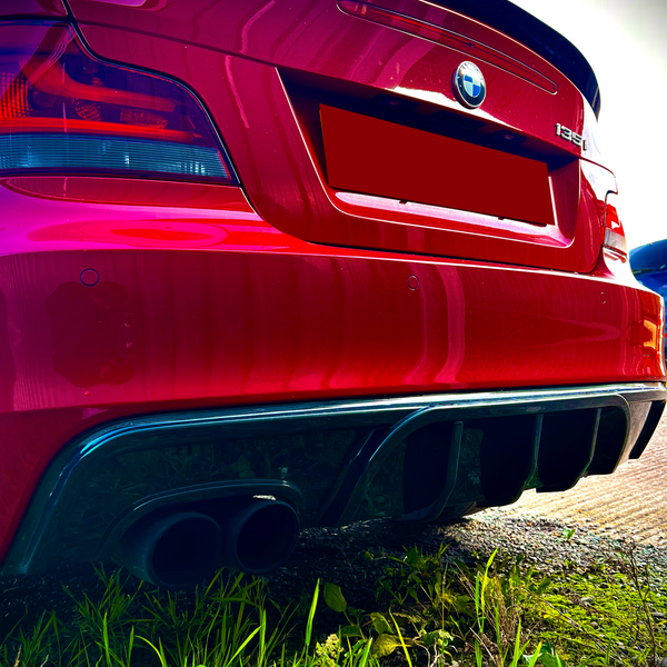 A red BMW 135i with dual exhaust pipes is parked on a grassy area. The concrete path is partially visible, and the rear of the car shows distinctive branding.