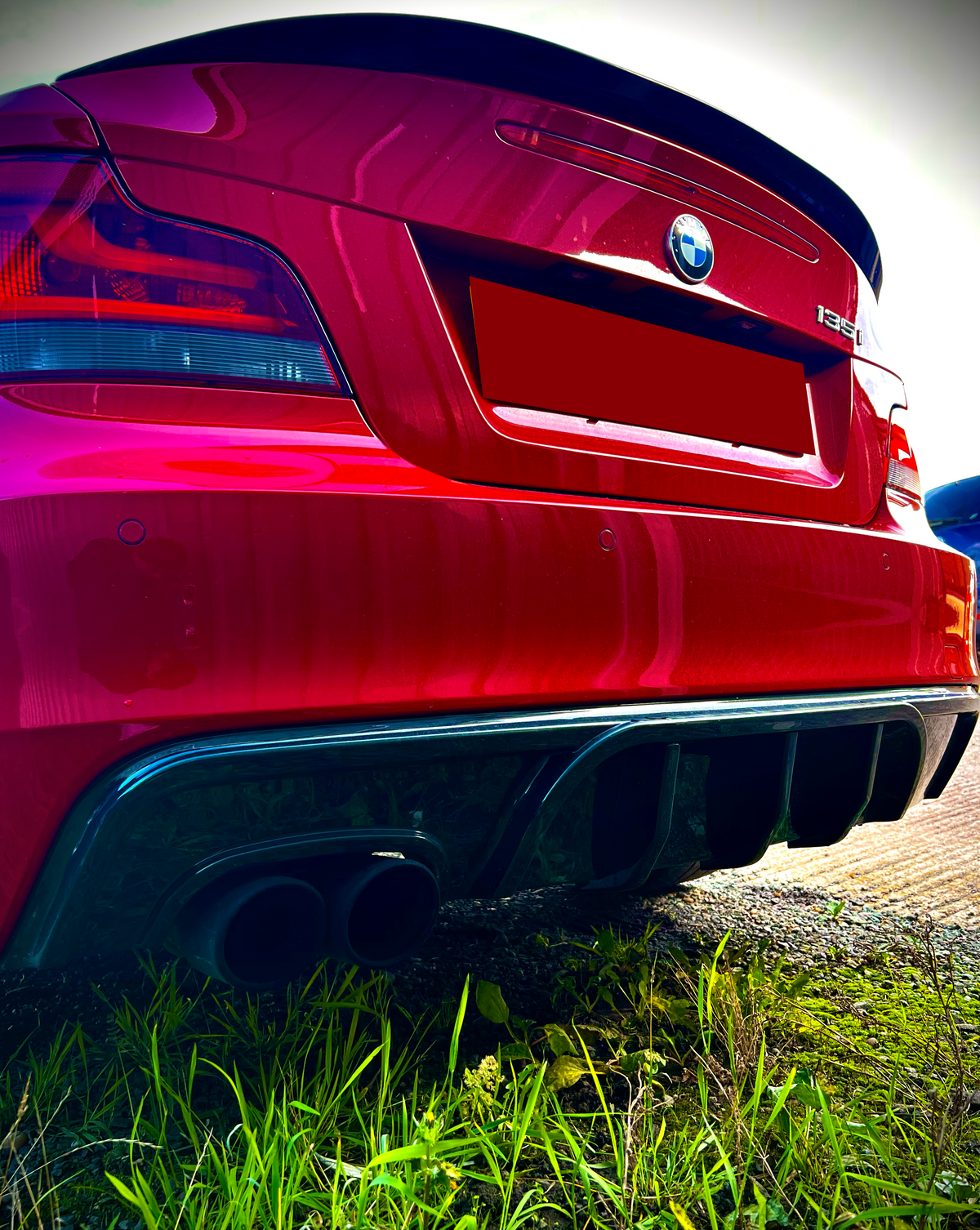 A red BMW 135i with dual exhaust pipes is parked on a grassy area. The concrete path is partially visible, and the rear of the car shows distinctive branding.