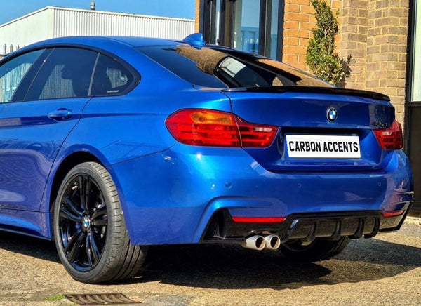 A blue BMW car is parked, showcasing its rear design and dual exhausts. The license plate reads "CARBON ACCENTS" in a sunny, urban setting with brick and glass structures.