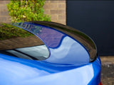 A blue car's rear spoiler reflects its surroundings, parked beside a brick wall and greenery, with a large, dark door in the background providing contrast.
