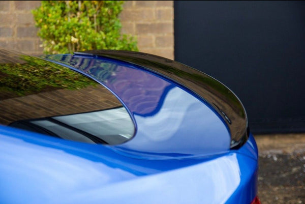 A blue car's rear spoiler reflects its surroundings, parked beside a brick wall and greenery, with a large, dark door in the background providing contrast.