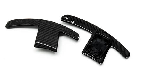 Two carbon-fiber paddle shifters, positioned side-by-side, lie on a plain white background. The right paddle features a plus sign and an arrow.
