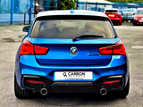 A blue BMW M140i is parked on an asphalt surface, surrounded by other vehicles. Text on the license plate reads: 
