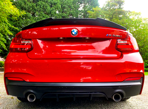 A red BMW M235i features prominently, parked on a gravel surface, surrounded by lush greenery. The BMW and M235i logos are visible on the back, with dual exhausts below.
