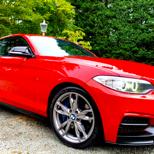 A red sports car, parked on a gravel driveway, is surrounded by lush green trees, with a classic lamp post in the background.