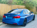A blue BMW 320d is parked on a paved street next to a wooden fence, surrounded by greenery and a 20 mph speed limit sign.
