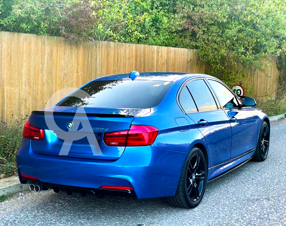 A blue BMW sedan is parked on a paved road, facing away, near a wooden fence with greenery. The model number "320d" is visible on the rear.