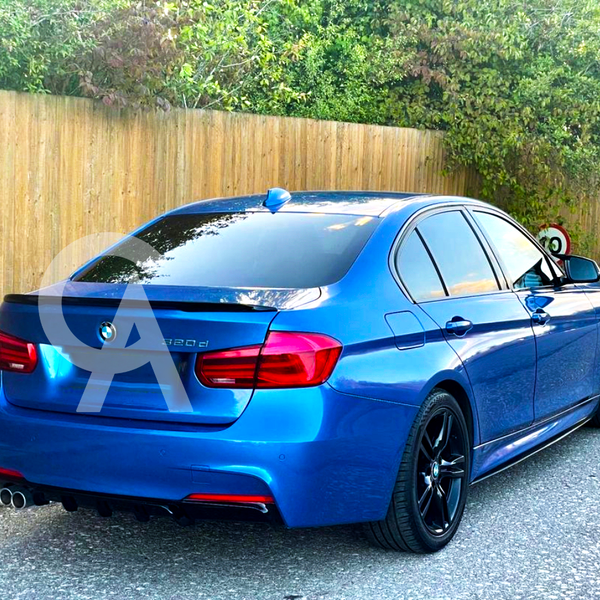 A blue BMW sedan is parked on a paved road, facing away, near a wooden fence with greenery. The model number "320d" is visible on the rear.
