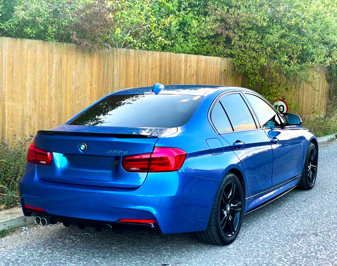 A blue BMW 320d is parked on a road, facing away. It features tinted windows and black wheels, adjacent to a wooden fence and greenery.