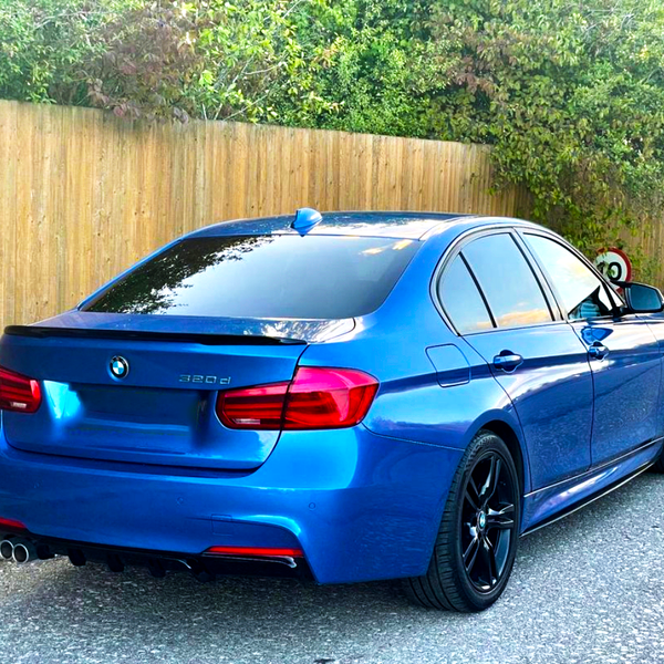 A blue BMW 320d is parked on a road, facing away. It features tinted windows and black wheels, adjacent to a wooden fence and greenery.