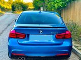 A blue BMW 320d is parked on a country road. Trees and a wooden fence line the roadside, creating a serene, rural environment.