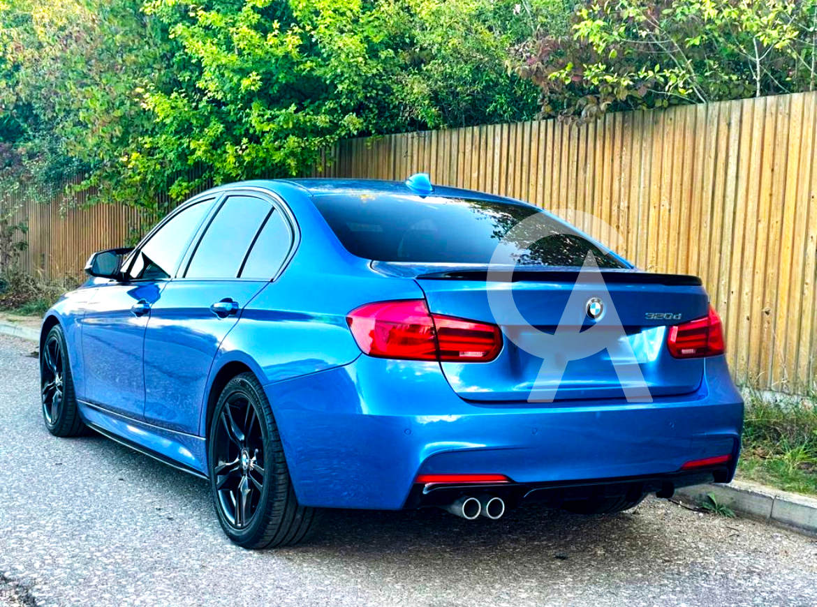 A blue BMW 320d car parked on a roadside, featuring tinted windows and dual exhaust pipes. A wooden fence and green foliage form the background.