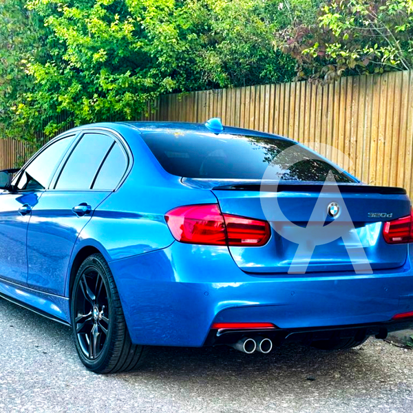 A blue BMW 320d car parked on a roadside, featuring tinted windows and dual exhaust pipes. A wooden fence and green foliage form the background.