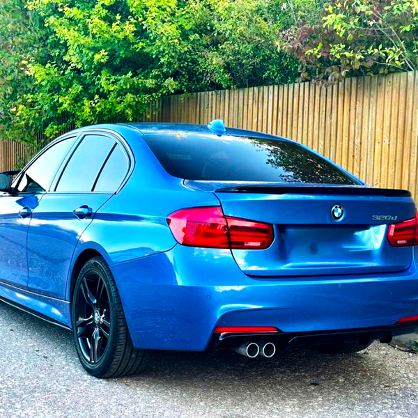 A blue BMW 320d car is parked on a street beside a wooden fence, surrounded by greenery. It features black wheels and dual exhaust pipes.