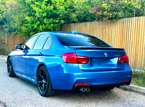 A blue BMW 320d car is parked on a street beside a wooden fence, surrounded by greenery. It features black wheels and dual exhaust pipes.