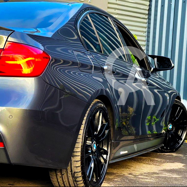 A sleek, grey sedan with tinted windows and bright red taillights is parked on pavement, reflecting striped patterns from a nearby corrugated metal wall and surrounded by green foliage.
