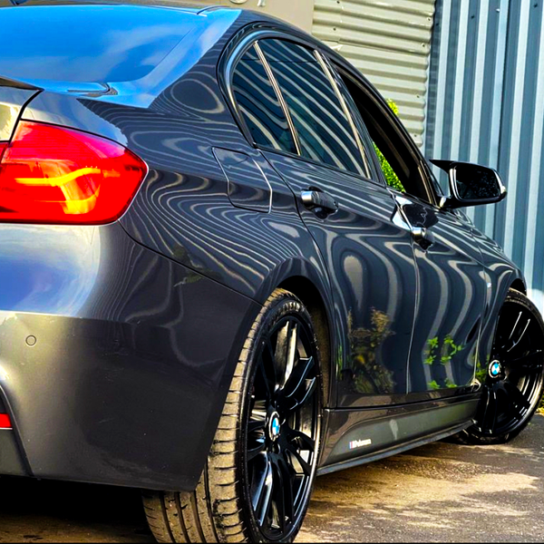 A sleek, dark gray car with illuminated red tail lights is parked on a paved surface, reflecting distorted patterns from a corrugated metal fence beside it.