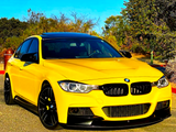 A yellow BMW car is parked, facing forward on a sunny driveway, surrounded by trees and shrubs under a clear blue sky.