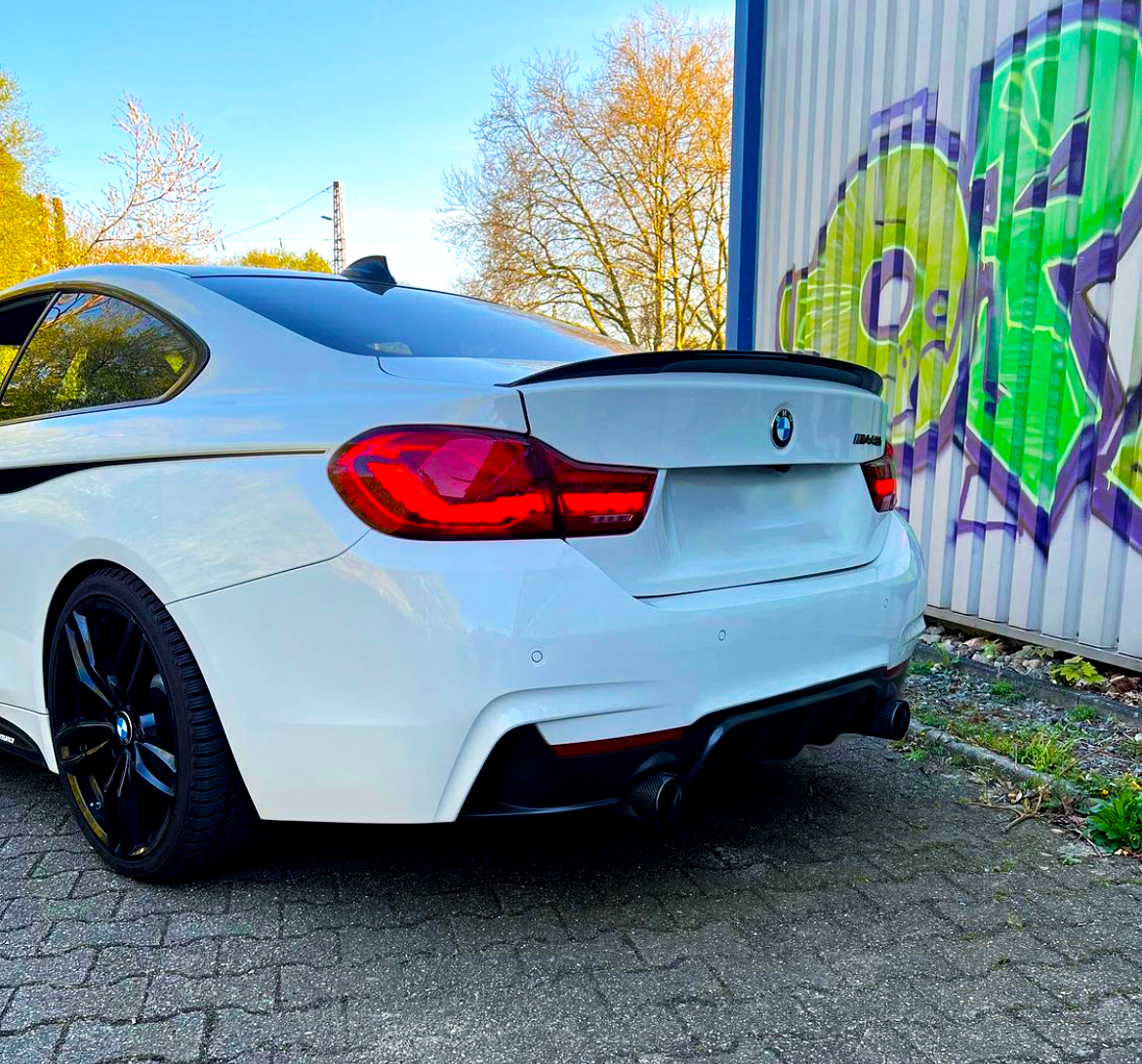 A white BMW parked on a cobblestone path near a wall with vibrant green and purple graffiti. Bare trees and a clear blue sky provide a crisp, open atmosphere.
