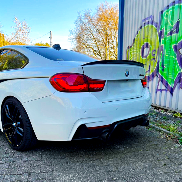 A white BMW parked on a cobblestone path near a wall with vibrant green and purple graffiti. Bare trees and a clear blue sky provide a crisp, open atmosphere.