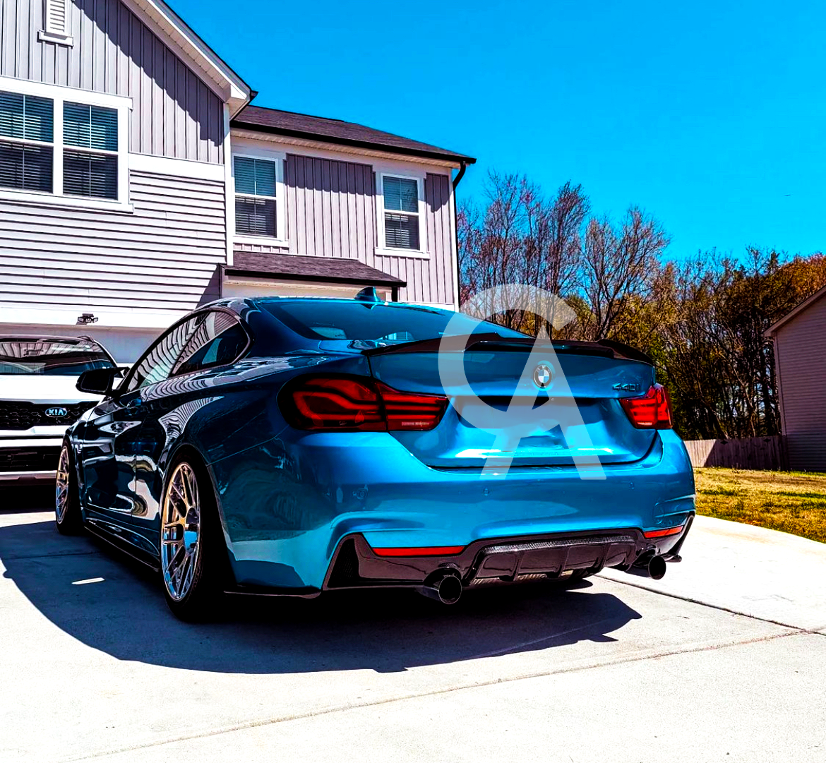 Blue BMW parked on a driveway, facing away from a two-story house. A white Kia is parked nearby, with trees and a blue sky in the background.