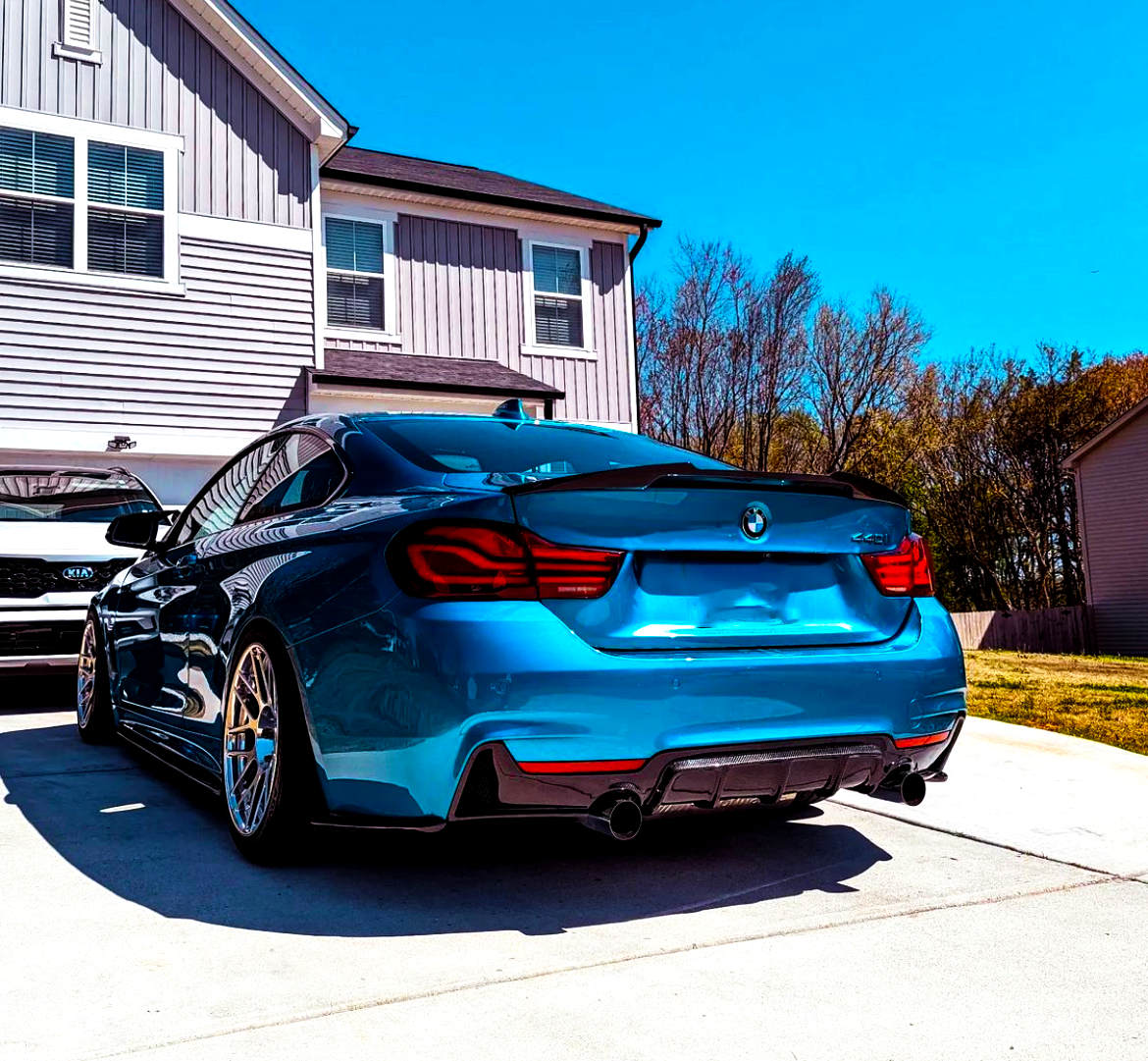 A blue BMW coupe is parked on a driveway beside a beige house. In the background, trees line the boundary, and a white Kia SUV is partially visible.