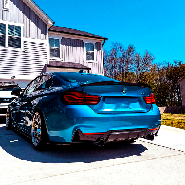 A blue BMW coupe is parked on a driveway beside a beige house. In the background, trees line the boundary, and a white Kia SUV is partially visible.