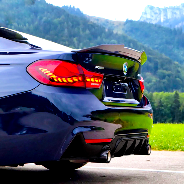 A sleek blue BMW car parked, highlighting its tail lights and exhaust system, with a forested mountain landscape in the background. Text: "XDrive 440i."