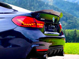 A dark blue BMW with illuminated taillights is parked on a paved road, surrounded by green fields and mountains. The car features dual exhausts. Visible text: 