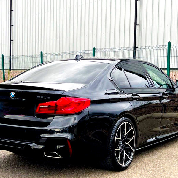 Black BMW sedan parked next to a fence, with shiny rims and distinctive red taillights, positioned on a concrete surface beside a large industrial-looking building.