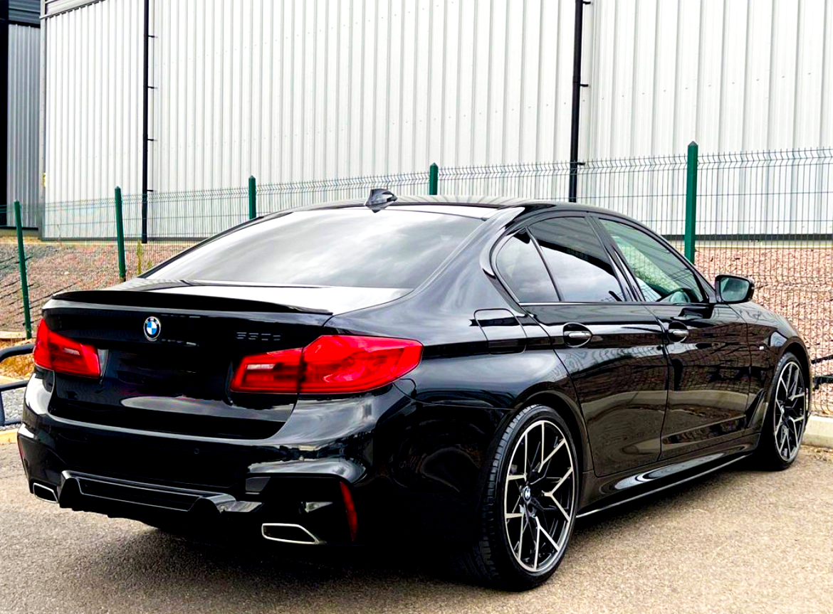 Black BMW sedan parked next to a fence, with shiny rims and distinctive red taillights, positioned on a concrete surface beside a large industrial-looking building.