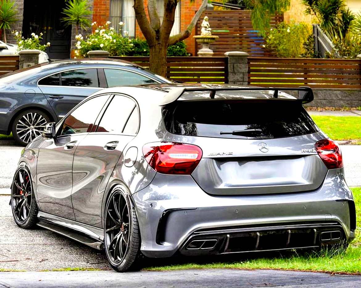 A silver sports hatchback with AMG and Mercedes badges, parked beside a road, features custom rims and a rear spoiler in a suburban neighborhood with trees and flowered gardens.