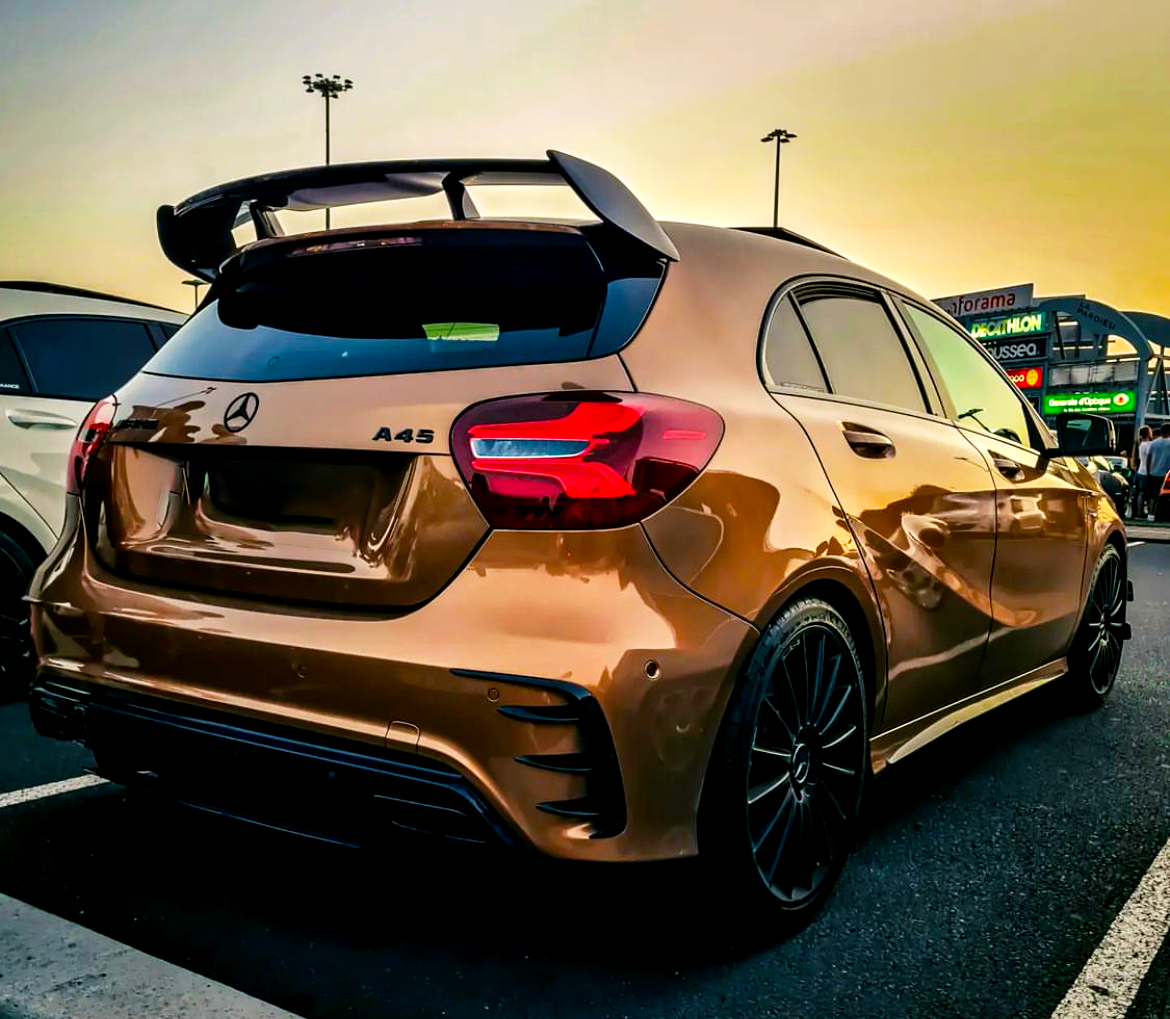 A bronze Mercedes A45 parked in a lot at sunset, featuring a prominent rear spoiler, black wheels, and illuminated taillights. Nearby, there are buildings with signs and people walking.