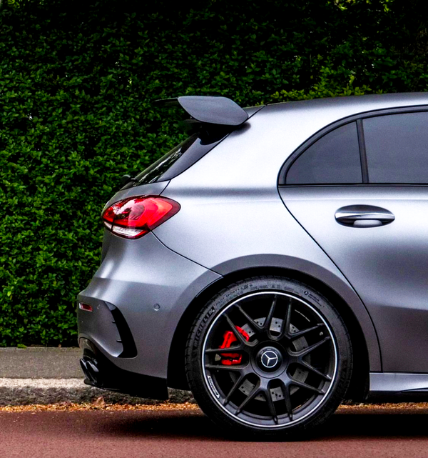 A silver car with a rear spoiler is parked beside a leafy hedge. Its red brake calipers are visible behind black alloy wheels.