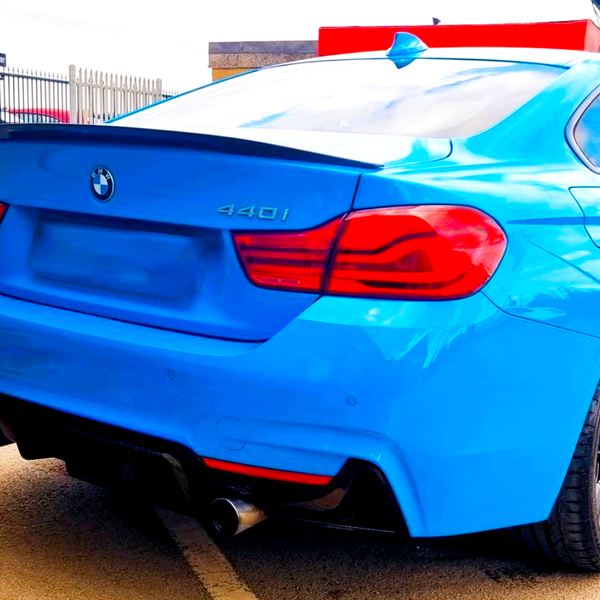 A blue BMW 440i car is parked on a light-colored surface, featuring dual exhausts and red-taillights. A fenced area and another parked car are visible in the background.