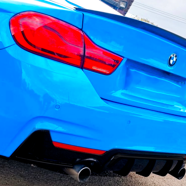 A blue BMW 440i with dual exhaust is parked on asphalt. Its distinctive rear lights and emblem are visible in bright daylight. Another car is partially visible nearby.