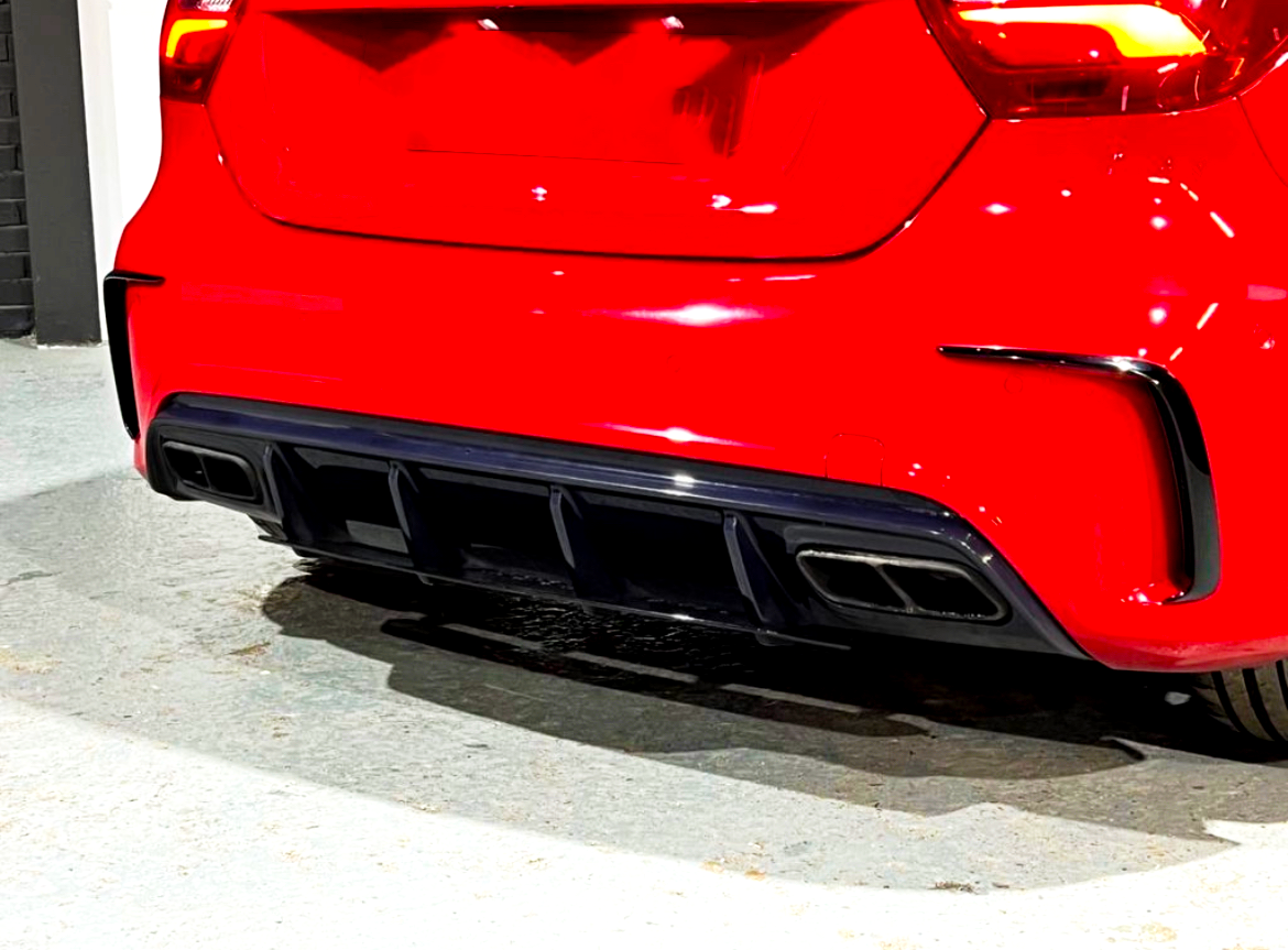 A red car with sleek taillights is parked, showcasing its rear bumper and dual exhaust outlets, on a concrete surface adjacent to a black and white wall.