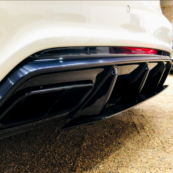 The image shows a car's white rear bumper with a black quad exhaust diffuser. It is parked indoors on a gravel surface, with another car partially visible in the background.