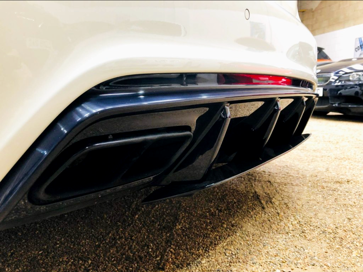 The image shows a car's white rear bumper with a black quad exhaust diffuser. It is parked indoors on a gravel surface, with another car partially visible in the background.