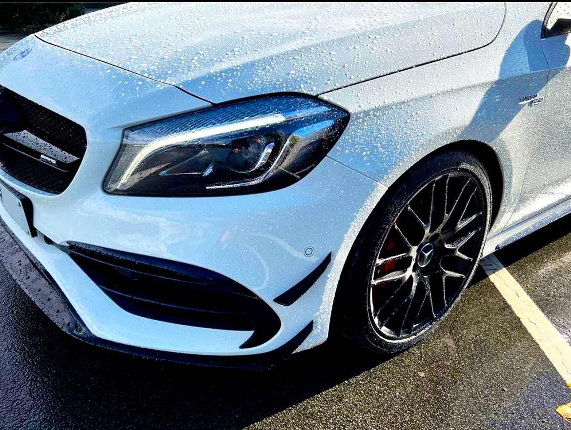 A white car, with water droplets on its surface, is parked in a lot. The vehicle features a sleek front design, prominent headlights, and shiny black alloy wheels.